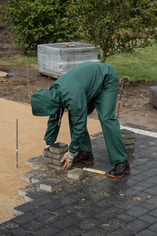 A person wearing green protective clothing and gloves is arranging square paving stones on a partially constructed pathway. The individual is bending over to pick up or place the bricks, which are stacked near their feet. The surroundings include a patch of grass, a wrapped pallet in the background, and a few trees.