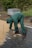 A person wearing green protective clothing and gloves is arranging square paving stones on a partially constructed pathway. The individual is bending over to pick up or place the bricks, which are stacked near their feet. The surroundings include a patch of grass, a wrapped pallet in the background, and a few trees.