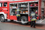 Two firefighters wearing uniforms with the word 'STRAZ' on the back are standing by the open side compartment of a red fire truck. The compartment is filled with various emergency response tools and equipment such as chainsaws, hoses, and mechanical devices. The ground is paved with bricks, and the scene appears to be a preparation or maintenance activity.