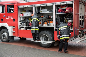 Two firefighters wearing uniforms with the word 'STRAZ' on the back are standing by the open side compartment of a red fire truck. The compartment is filled with various emergency response tools and equipment such as chainsaws, hoses, and mechanical devices. The ground is paved with bricks, and the scene appears to be a preparation or maintenance activity.
