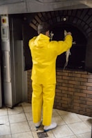 A person wearing a bright yellow rain suit is holding fish in front of an industrial oven. The surroundings have a tiled floor and brickwork under the oven.