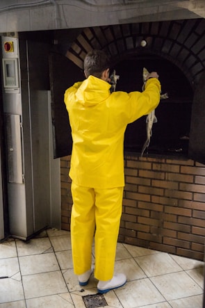 A person wearing a bright yellow rain suit is holding fish in front of an industrial oven. The surroundings have a tiled floor and brickwork under the oven.