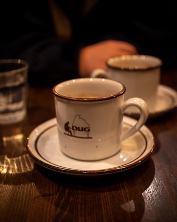 A close-up of a white coffee cup with a dark rim sits on a saucer on a wooden table. The cup features a logo with the word 'DUG' and an illustration of a person playing a grand piano. Another similar cup is partially visible in the background, along with a glass of water. A person's arm is resting on the table, adding a casual human element.