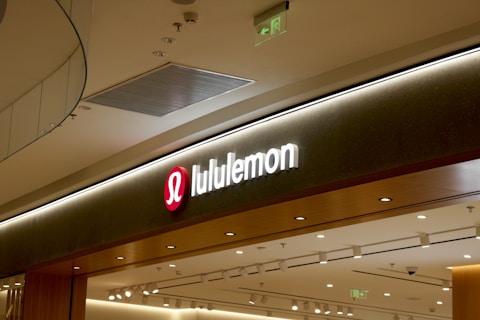 A retail store sign with the brand name 'lululemon' illuminated in white letters on a dark background. The sign includes the brand's recognizable logo, a stylized 'A' in a red circle to the left of the name. The store's interior features modern lighting and design elements with a ceiling composed of recessed lighting and metallic ventilation grates.
