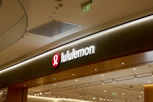 A retail store sign with the brand name 'lululemon' illuminated in white letters on a dark background. The sign includes the brand's recognizable logo, a stylized 'A' in a red circle to the left of the name. The store's interior features modern lighting and design elements with a ceiling composed of recessed lighting and metallic ventilation grates.