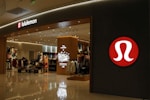 A well-lit retail store featuring a modern and sleek entrance with prominent branding. The interior is visible with displays of clothing and accessories, suggesting an apparel store. The store has a structured layout and several mannequins.
