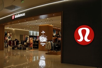 A well-lit retail store featuring a modern and sleek entrance with prominent branding. The interior is visible with displays of clothing and accessories, suggesting an apparel store. The store has a structured layout and several mannequins.