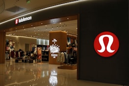 A well-lit retail store featuring a modern and sleek entrance with prominent branding. The interior is visible with displays of clothing and accessories, suggesting an apparel store. The store has a structured layout and several mannequins.
