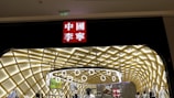A storefront with an illuminated interior featuring a modern, geometric ceiling design. Numerous mannequins display clothing, and a prominent red sign with white Chinese characters is situated above the entrance.
