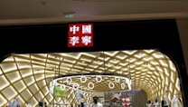 A storefront with an illuminated interior featuring a modern, geometric ceiling design. Numerous mannequins display clothing, and a prominent red sign with white Chinese characters is situated above the entrance.