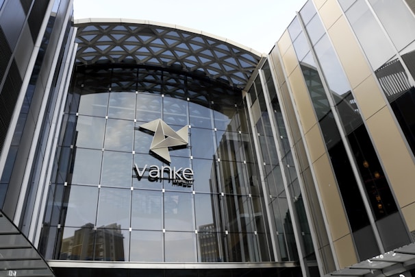 A modern glass building with a geometric pattern on the roof and a large logo in the center. The facade is mainly composed of glass panels with metal frames, reflecting some nearby structures.