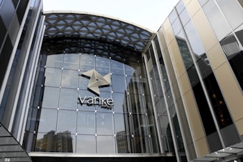 A modern glass building with a geometric pattern on the roof and a large logo in the center. The facade is mainly composed of glass panels with metal frames, reflecting some nearby structures.