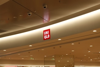 The interior of a modern shopping mall features a prominent Uniqlo store sign displayed on a light-colored ceiling. The red Uniqlo logo stands out against the neutral tones of the surroundings, which include recessed lighting and surveillance cameras.
