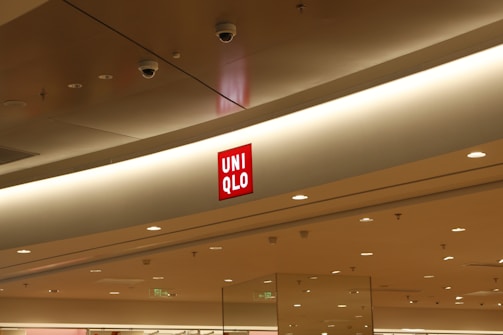 The interior of a modern shopping mall features a prominent Uniqlo store sign displayed on a light-colored ceiling. The red Uniqlo logo stands out against the neutral tones of the surroundings, which include recessed lighting and surveillance cameras.