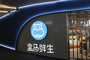 A storefront of a retail location featuring a large, prominently displayed logo of a stylized blue hippo. The logo is circular and placed on a dark panel. Below the logo, there are Chinese characters lit in white. Inside the store, visible through large windows, are shoppers and various grocery items.