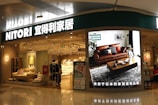A store entrance with a prominent sign reading 'NITORI' in Latin and Chinese characters. The store has a modern interior displaying furniture items such as sofas and cushions. A large digital screen near the entrance showcases a living room setting with a brown leather sofa, a coffee table, and decorative plants. The store is brightly lit, and a few people are visible walking inside.