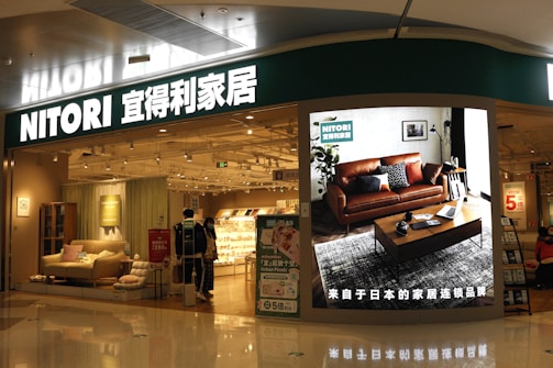 A store entrance with a prominent sign reading 'NITORI' in Latin and Chinese characters. The store has a modern interior displaying furniture items such as sofas and cushions. A large digital screen near the entrance showcases a living room setting with a brown leather sofa, a coffee table, and decorative plants. The store is brightly lit, and a few people are visible walking inside.