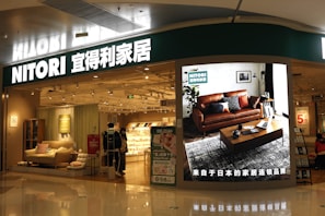 A store entrance with a prominent sign reading 'NITORI' in Latin and Chinese characters. The store has a modern interior displaying furniture items such as sofas and cushions. A large digital screen near the entrance showcases a living room setting with a brown leather sofa, a coffee table, and decorative plants. The store is brightly lit, and a few people are visible walking inside.