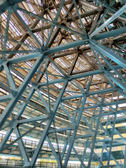 Metallic beams and trusses form an intricate lattice structure. The framework consists of intersecting steel beams, creating a geometric pattern with an industrial aesthetic. Light filters through gaps in the structure, casting shadows and highlighting the engineering details.