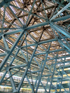 Metallic beams and trusses form an intricate lattice structure. The framework consists of intersecting steel beams, creating a geometric pattern with an industrial aesthetic. Light filters through gaps in the structure, casting shadows and highlighting the engineering details.