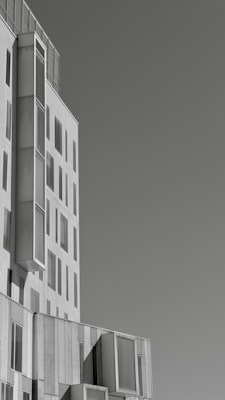 A modern, multi-story building with a minimalist design, featuring large, rectangular windows and a light, monochromatic façade. The architecture includes a series of protruding sections, giving the structure depth and an intriguing geometric form.