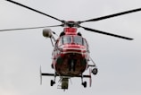 A red helicopter is captured mid-flight against a muted, cloudy sky. The helicopter's rotors are spinning, and various components such as landing skids and cabin details are visible.