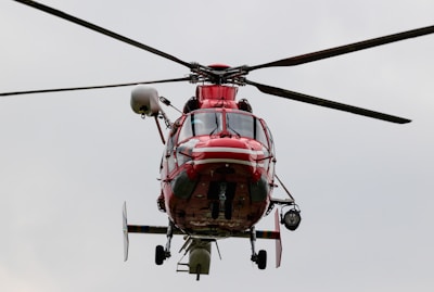 A red helicopter is captured mid-flight against a muted, cloudy sky. The helicopter's rotors are spinning, and various components such as landing skids and cabin details are visible.