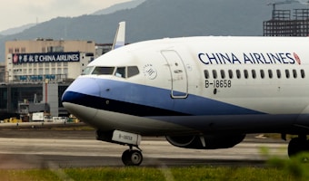 An airplane with the branding of China Airlines is positioned on an airport tarmac. The aircraft is a commercial passenger jet, featuring a white and blue color scheme with the registration number B-18658 visible. In the background, there is a building with signage for China Airlines, indicating an airport setting with mountainous terrain in the distance.