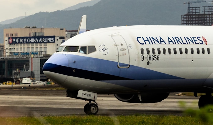 An airplane with the branding of China Airlines is positioned on an airport tarmac. The aircraft is a commercial passenger jet, featuring a white and blue color scheme with the registration number B-18658 visible. In the background, there is a building with signage for China Airlines, indicating an airport setting with mountainous terrain in the distance.