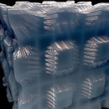 A close-up view of a textured, translucent material with a geometric pattern. The surface appears smooth with wavy, rippling lines running across it, reflecting light in a dynamic way. The structure is three-dimensional, resembling a cube or block with intricate details.