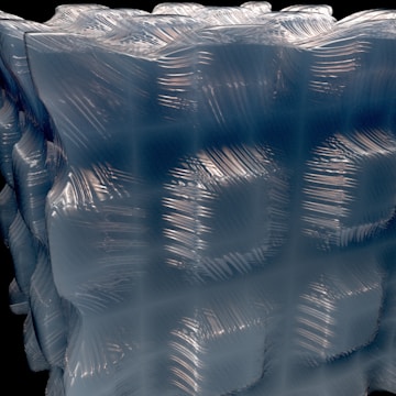 A close-up view of a textured, translucent material with a geometric pattern. The surface appears smooth with wavy, rippling lines running across it, reflecting light in a dynamic way. The structure is three-dimensional, resembling a cube or block with intricate details.