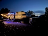 A travel trailer is parked at a campsite during twilight. The trailer is illuminated by a string of warm, glowing lights hanging from an extended awning. Several outdoor chairs and a table are positioned on a rug next to the trailer, creating a cozy outdoor seating area. A truck is parked adjacent to the trailer. Trees and a darkening sky provide a tranquil backdrop.