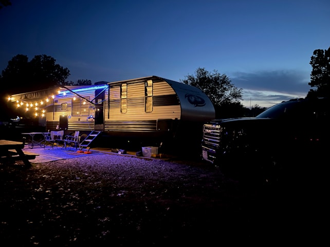 A travel trailer is parked at a campsite during twilight. The trailer is illuminated by a string of warm, glowing lights hanging from an extended awning. Several outdoor chairs and a table are positioned on a rug next to the trailer, creating a cozy outdoor seating area. A truck is parked adjacent to the trailer. Trees and a darkening sky provide a tranquil backdrop.