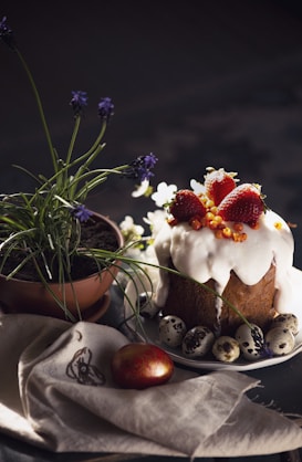 A festive table setting featuring a beautifully decorated cake topped with glossy icing, strawberries, and small orange berries. Surrounding the cake are speckled eggs, a potted plant with purple flowers, and a shiny decorated egg resting on a cloth.