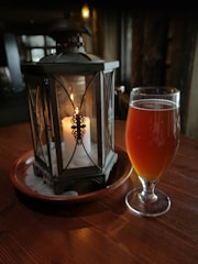 A glass of amber-colored beverage is placed on a wooden table beside an ornate metal lantern with a lit candle inside. The setting appears dimly lit, creating a warm and cozy ambiance.