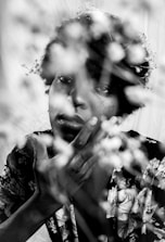 A black and white photograph features a person with a thoughtful expression, their hand gently touching their chin. Delicate flowers partially obscure the view in the foreground, creating a soft and dreamy effect.