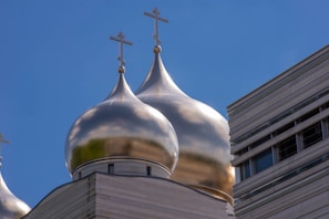 Golden onion domes with crosses adorn the top of a building against a clear blue sky. The architecture on the left appears traditional, while the structure on the right has a modern design with horizontal lines and windows.