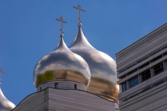 Golden onion domes with crosses adorn the top of a building against a clear blue sky. The architecture on the left appears traditional, while the structure on the right has a modern design with horizontal lines and windows.