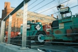 The image depicts a construction site featuring machinery and equipment surrounded by metal railings. In the background, an old brick building with rounded towers and several windows is visible. The foreground includes a green machine labeled with a 'HOT 3257' sign and a red cylindrical object. The scene is bathed in natural daylight, casting soft shadows.