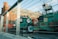 The image depicts a construction site featuring machinery and equipment surrounded by metal railings. In the background, an old brick building with rounded towers and several windows is visible. The foreground includes a green machine labeled with a 'HOT 3257' sign and a red cylindrical object. The scene is bathed in natural daylight, casting soft shadows.