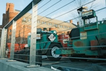 The image depicts a construction site featuring machinery and equipment surrounded by metal railings. In the background, an old brick building with rounded towers and several windows is visible. The foreground includes a green machine labeled with a 'HOT 3257' sign and a red cylindrical object. The scene is bathed in natural daylight, casting soft shadows.