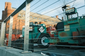 The image depicts a construction site featuring machinery and equipment surrounded by metal railings. In the background, an old brick building with rounded towers and several windows is visible. The foreground includes a green machine labeled with a 'HOT 3257' sign and a red cylindrical object. The scene is bathed in natural daylight, casting soft shadows.