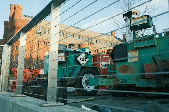 The image depicts a construction site featuring machinery and equipment surrounded by metal railings. In the background, an old brick building with rounded towers and several windows is visible. The foreground includes a green machine labeled with a 'HOT 3257' sign and a red cylindrical object. The scene is bathed in natural daylight, casting soft shadows.