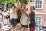 A group of four people is gathered outside, toasting with drinks under the shade of trees. They are smiling and appear to be enjoying a social gathering. There is a building with a brick wall and a rainbow flag hanging in the background. Other people are visible behind them, indicating a lively event.