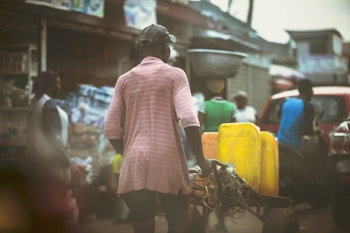 A man wearing a pink shirt and cap is pushing a cart with yellow jerrycans along a busy street market. There are multiple people in the background, some standing near stalls filled with various goods. The image is slightly desaturated, giving it a vintage feel.