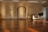 A spacious room with wooden flooring and warm lighting. There are several tripods with microphones and a spotlight on stands positioned around the room. A wooden archway is featured prominently on the back wall. A desk with a cut of fabric on top and a folded chair beside it is positioned on one side. The room appears to have an orderly and prepared setup, possibly for an event or presentation.