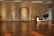 A spacious room with wooden flooring and warm lighting. There are several tripods with microphones and a spotlight on stands positioned around the room. A wooden archway is featured prominently on the back wall. A desk with a cut of fabric on top and a folded chair beside it is positioned on one side. The room appears to have an orderly and prepared setup, possibly for an event or presentation.