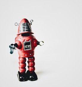 A vintage-style red robot toy with metallic accents stands against a plain white background. The robot features a dome-shaped head with antennae, claw-like hands, and a square display on its chest. Its legs are segmented and it has a wind-up key on its side.