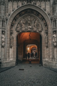 An ornate architectural entrance featuring detailed stone carvings of figures on the archway. The passageway is illuminated with warm light, creating a welcoming atmosphere as a few people walk through, blending historical craftsmanship with a bustling modern environment.