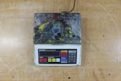 A digital scale with a keypad and display screen is covered in multicolored paint splatters, resting on a wooden surface.